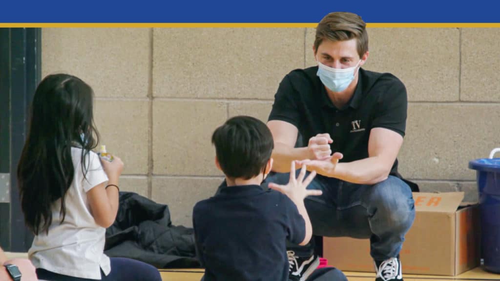 A man focusing on youth leadership sits in front of a young boy and girl and plays rock, paper, scissors with them.