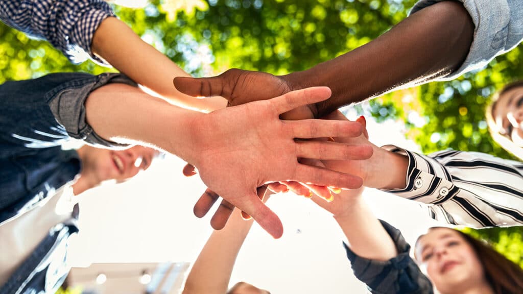 A group of young people participating in community action gather in a circle, their hands stacked in the middle